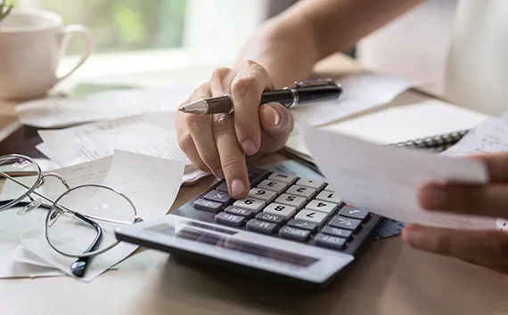 Person using calculator at desk with documents, glasses, and coffee cup visible during financial work or accounting task