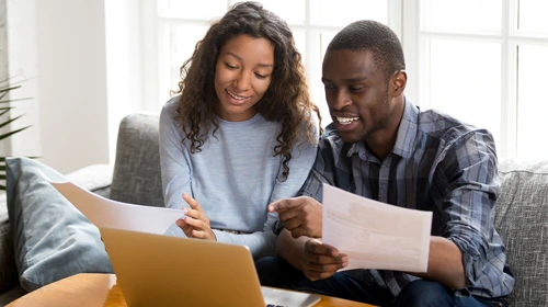 A man and woman sitting on a couch reviewing documents together at a laptop, smiling while discussing paperwork in a bright home office setting.