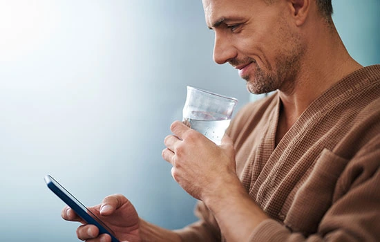 Man in brown shirt holding glass of water while looking at smartphone