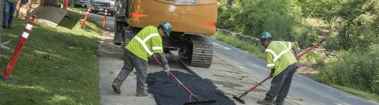 Two workers in yellow safety vests operating asphalt paving equipment on a residential driveway during road construction work