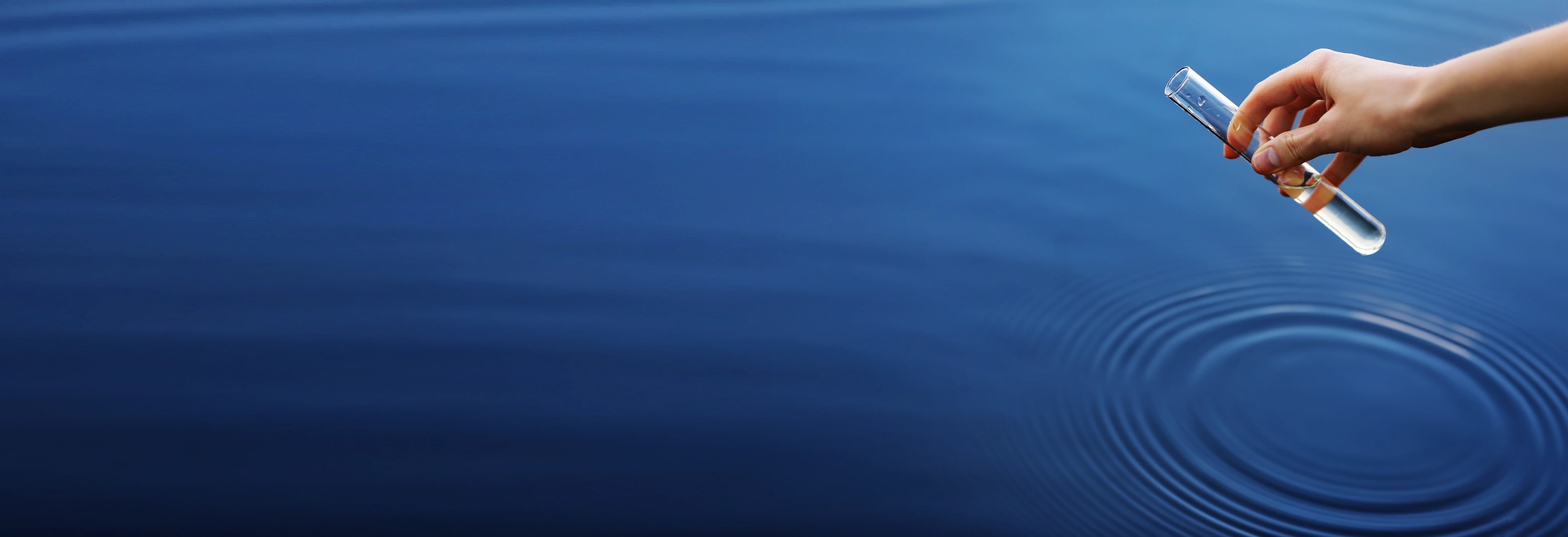 Hand dropping a water droplet into calm blue water creating concentric ripples on the surface